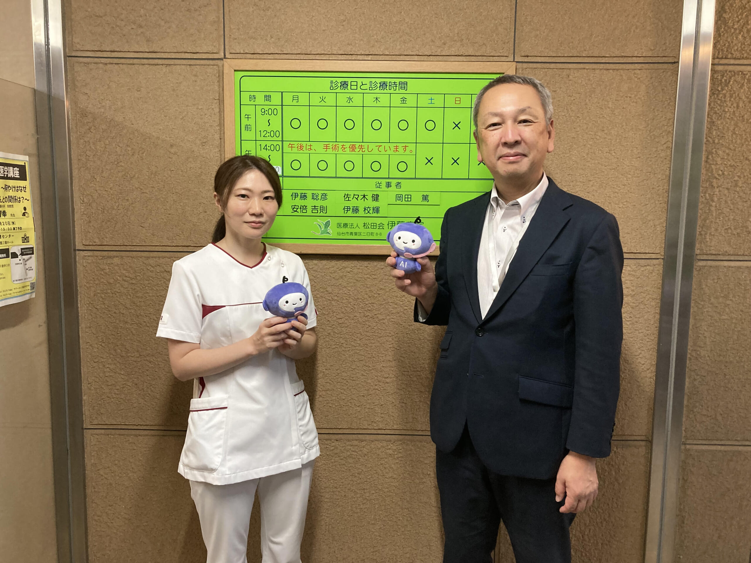 医療法人松田会 伊藤病院の看板前。左側が副看護師長の小菅さん、右側が事務部の佐竹さん